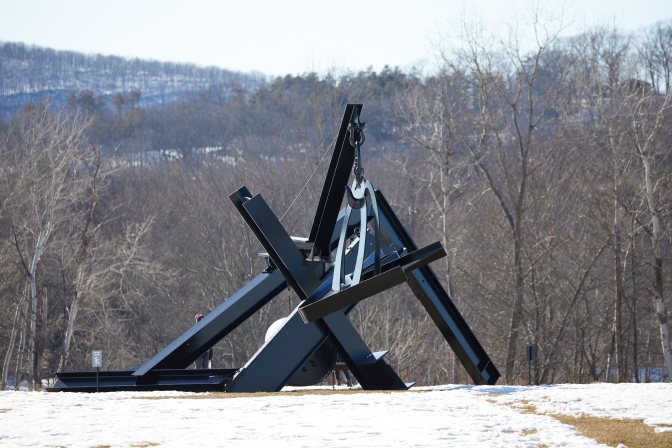 Metal sculpture with wooden bed hanging from crane arm.