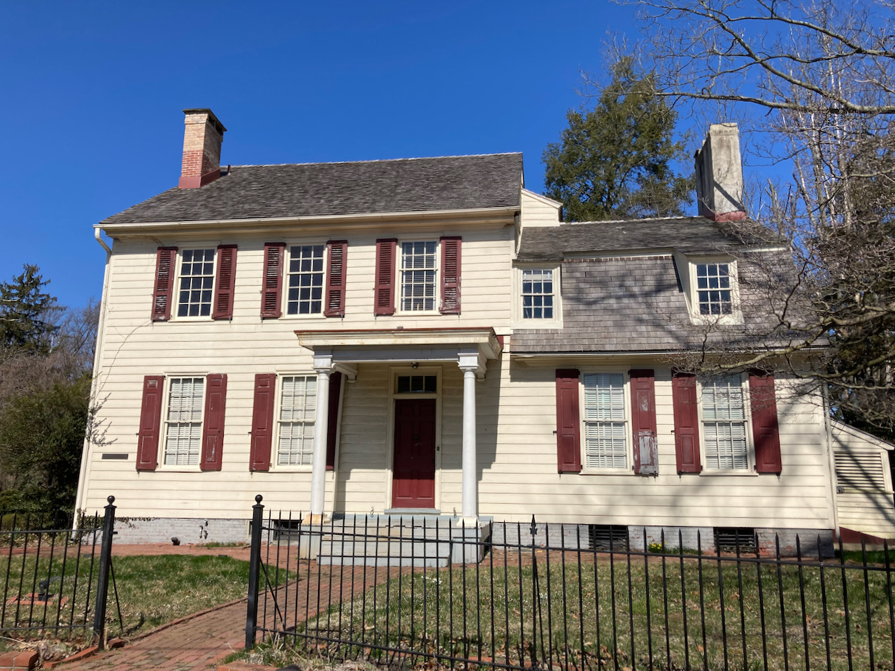 Tan two-story house, with two chimneys, surrounded by a wrought iron fence.