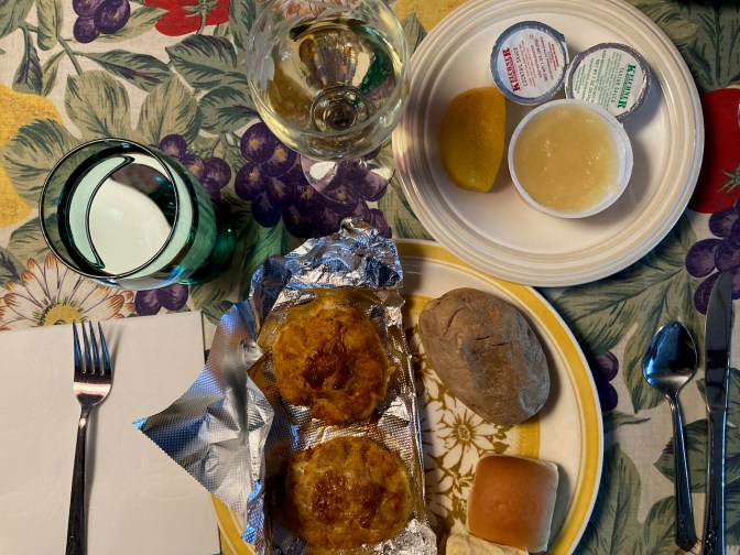 Plate with two crab cakes, baked potato, and roll, a glass of water, and a glass of wine.