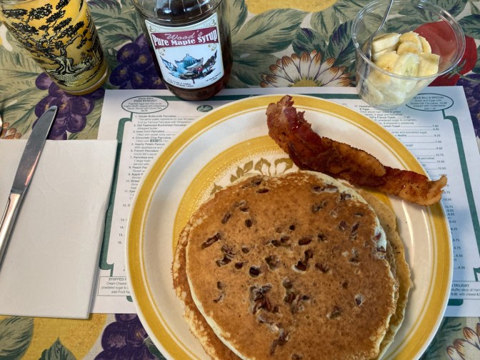 Pecan pancakes and bacon on a plate, with a glass of orange juice and a cup of cut bananas on table.