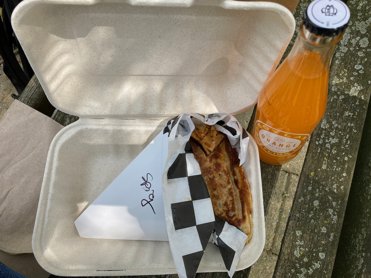 Crepe in paper wrapper, in paper box, with bottle of orange soda beside it.