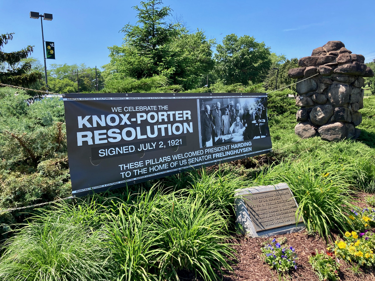 Small stone marker with plaque amid shrubbery. A large sign is hanging in the background, that states WE CELEBRATE THE KNOX-PORTER RESOLUTION SIGNED JULY 2, 1921 - THESE PILLARES WELCOMES PRESIDENT HARDING TO TEH HOME OF US SENATOR FRELINGHUYSEN.