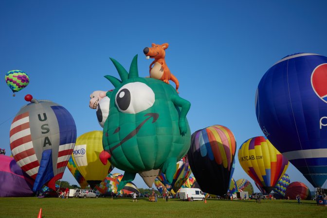 Balloons on ground, ready to take off, including one shaped as a green monster, and another shaped as a rocket ship with an American flag on it.