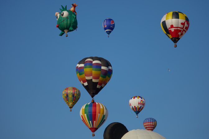 Mass ascension of several multicolored balloons, with green monster balloon in background.