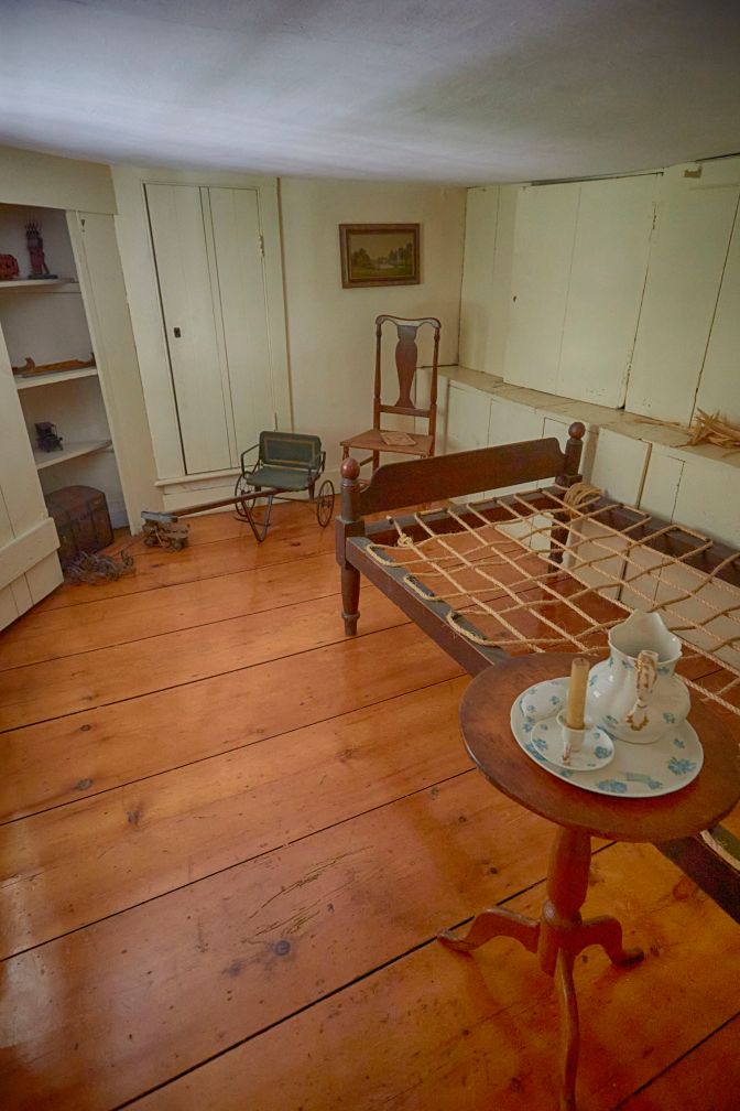 Room with low ceiling, rope bed frame, side table, and small door in back.
