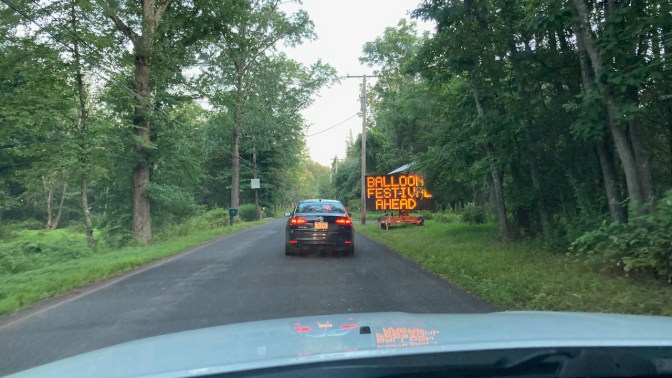 View of two-lane road with traffic, and a sign on right that says BALLOON FESTIVAL AHEAD.