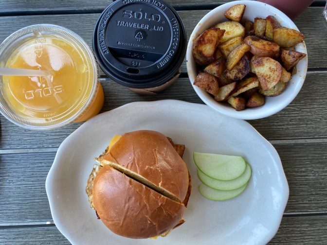 Breakfast sandwich on plate, with bowl of home fries, cup of orange juice, and cup of coffee.