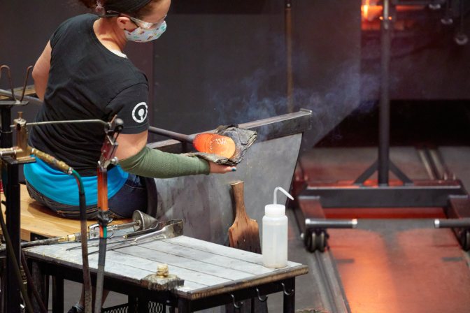 Glass worker, with hot glass on blow pipe, shaping glass with pad in hand.