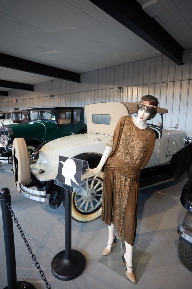 Mannequin of woman, in flapper dress, beside classic cars.