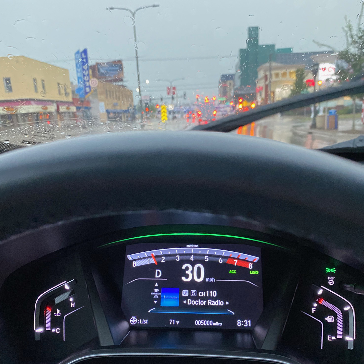 View of rainy street from behind windshield of vehicle. Dashboard in front has odometer that reads 5000 miles.