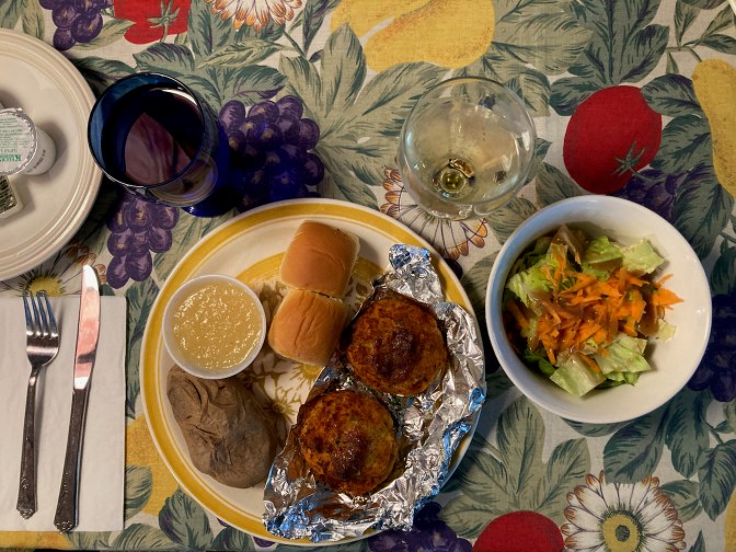 Plate with rolls, baked potato, apple sauce, and crab cakes, with a bowl with salad, a glass of wine, and a glass of water.