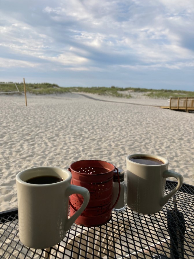 Two cups of coffee on metal table with beach in background.