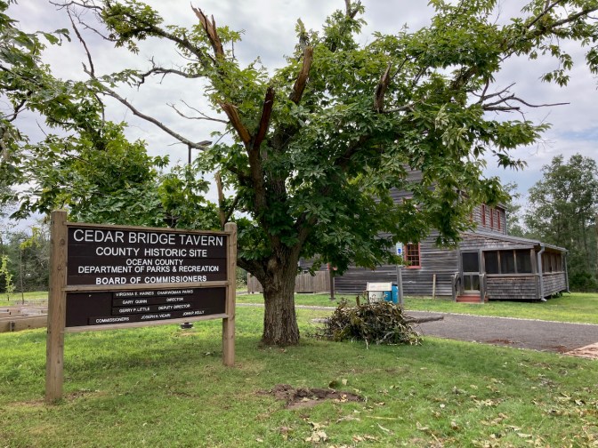 Sign that says Cedar Bridge Tavern - County Historic Site - Ocean County Board of Parks and Recreation, with a large building behind it.