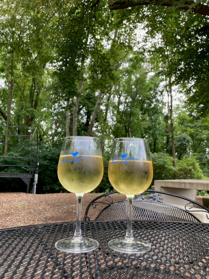 Two glasses of white wine on table, with trees in background.