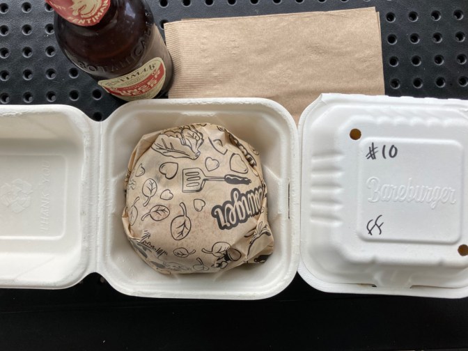 Burger in wax paper wrapping, box with fries, and a ginger beer bottle on metal table.