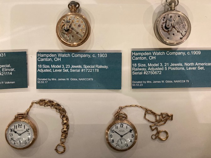 Four pocket watches in display case, two front-facing and two with their rear movements opened.