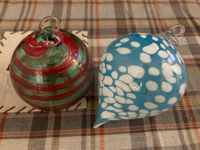 Two Christmas ornaments, one blue and white, one green and red, on plaid tablecloth.