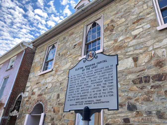Exterior of stone building, with sign in front telling history of quick wedding chapels in Elkton.