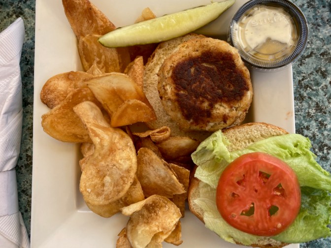 Crab cake sandwich on bun, with potato chips and pickle, on white plate.