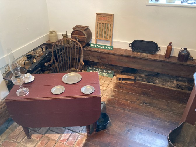 Kitchen, with two different floor levels - 1827 and 1884. Kitchen furnishings and a small dining table are in the room.