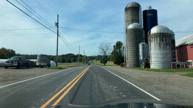 Farm with silos on right side of road, along two-lane country road. 