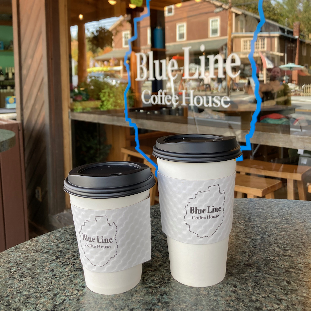 Two coffee cups on table in front of window. Window has Blue Line Coffee House decal.