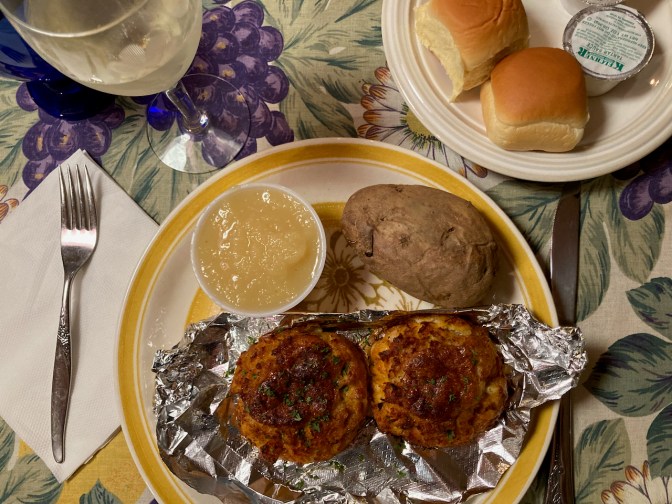Two crab cakes on plate, with baked potato and applesauce. 
