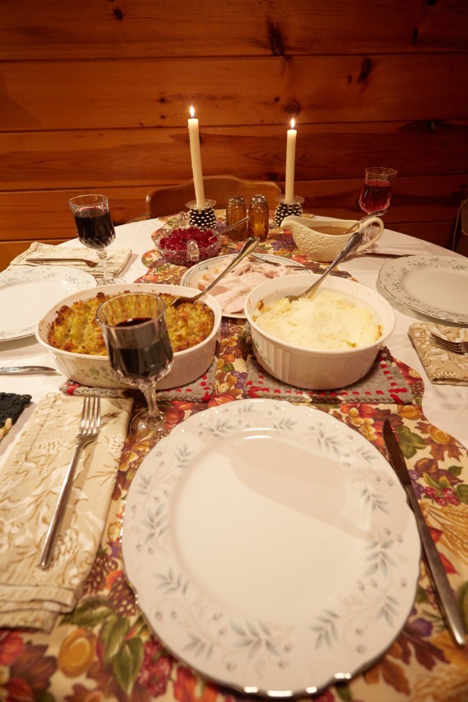 Table, with plates, bowl of mashed potatoes, bowl of stuffing, plate of turkey, gravy in gravy boat, cranberry sauce in bowl, three glasses of wine, and two candles. 
