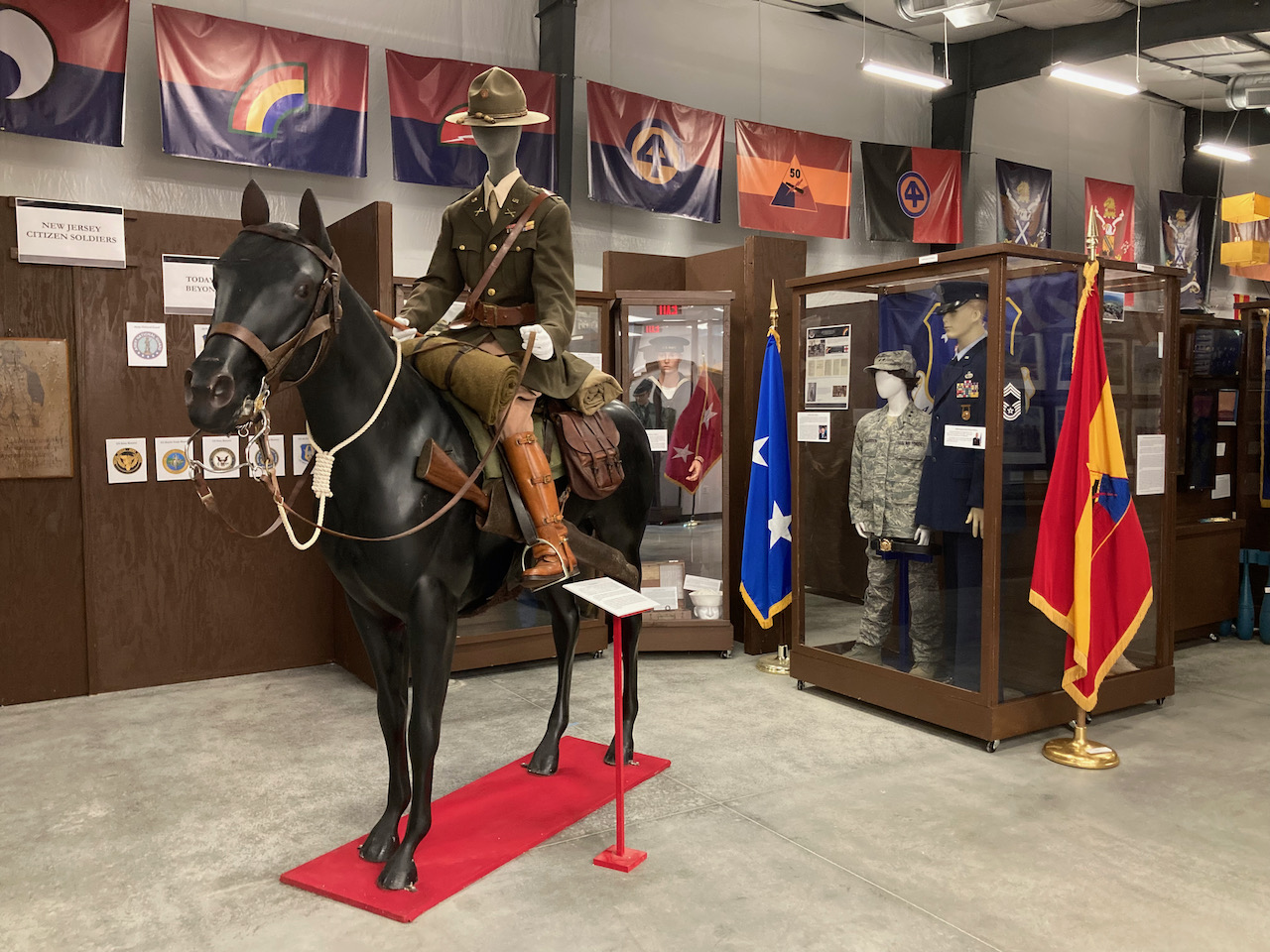 Mannequin of soldier riding on a horse. Flags of units hang from the ceiling, and mannequins of soldiers are in the background in display cases. 