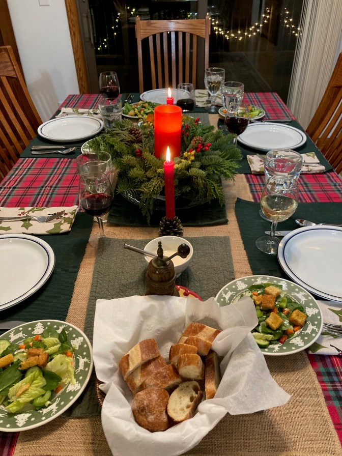 Dinner table set for Christmas, with a wreath and candle in center, plates at five places, and salads, bread, and glasses of water and wine.