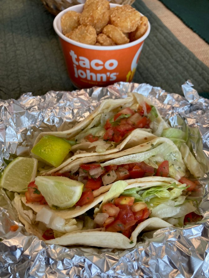 Three street tacos on aluminum foil, with basket of tater tots in background.