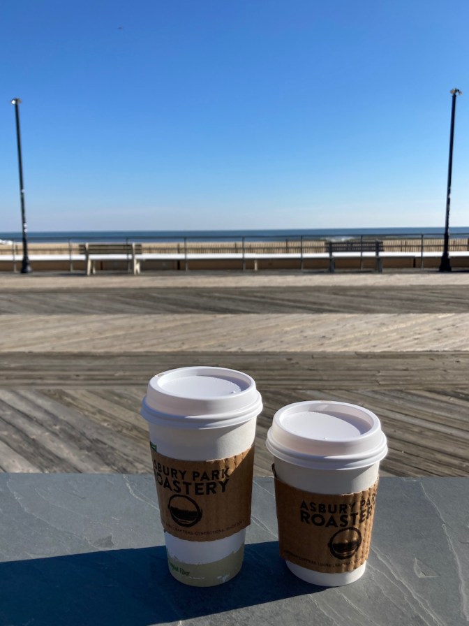 Two cups of coffee in foreground, with boardwalk and ocean in background.