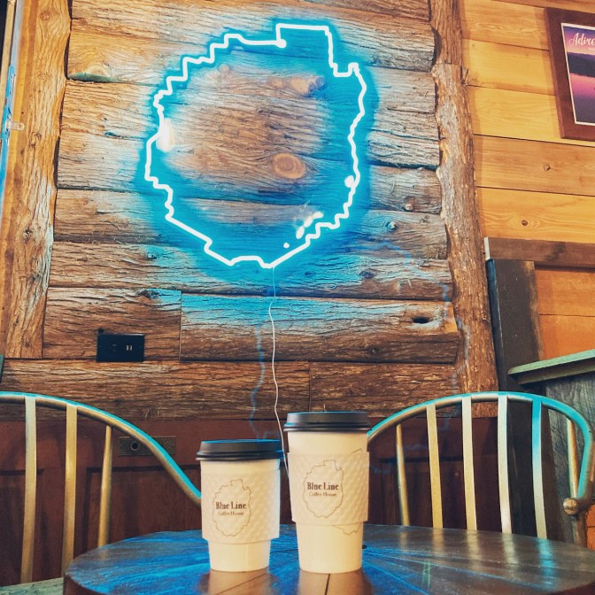 Two cups of coffee on table, with neon sign outline of Adirondack State Park in background.