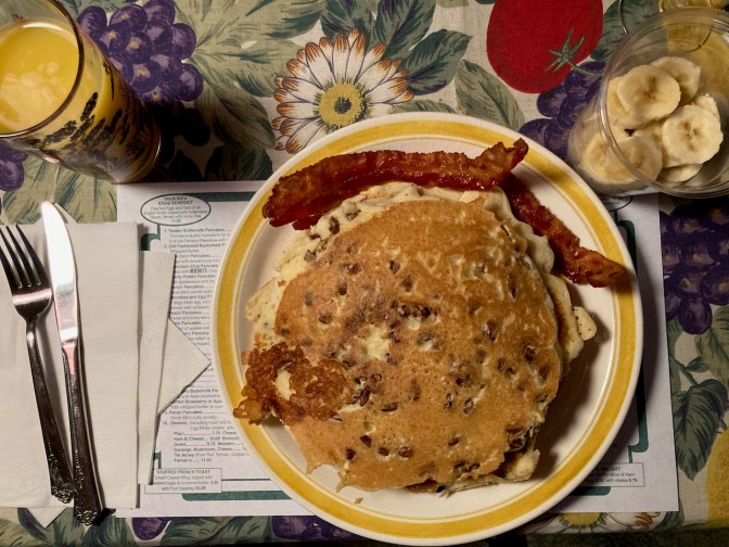 Plate with pancakes and bacon, small cup of bananas, and glass of orange juice.