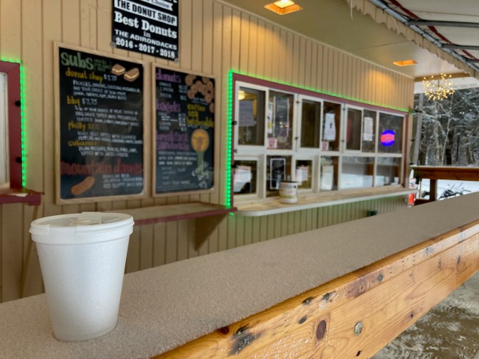 Cup of coffee on banister with ordering window of donut shop in background.
