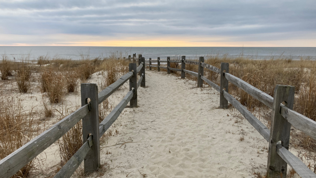 View of sandy path over dune leading to beach, with grey sky over ocean.
