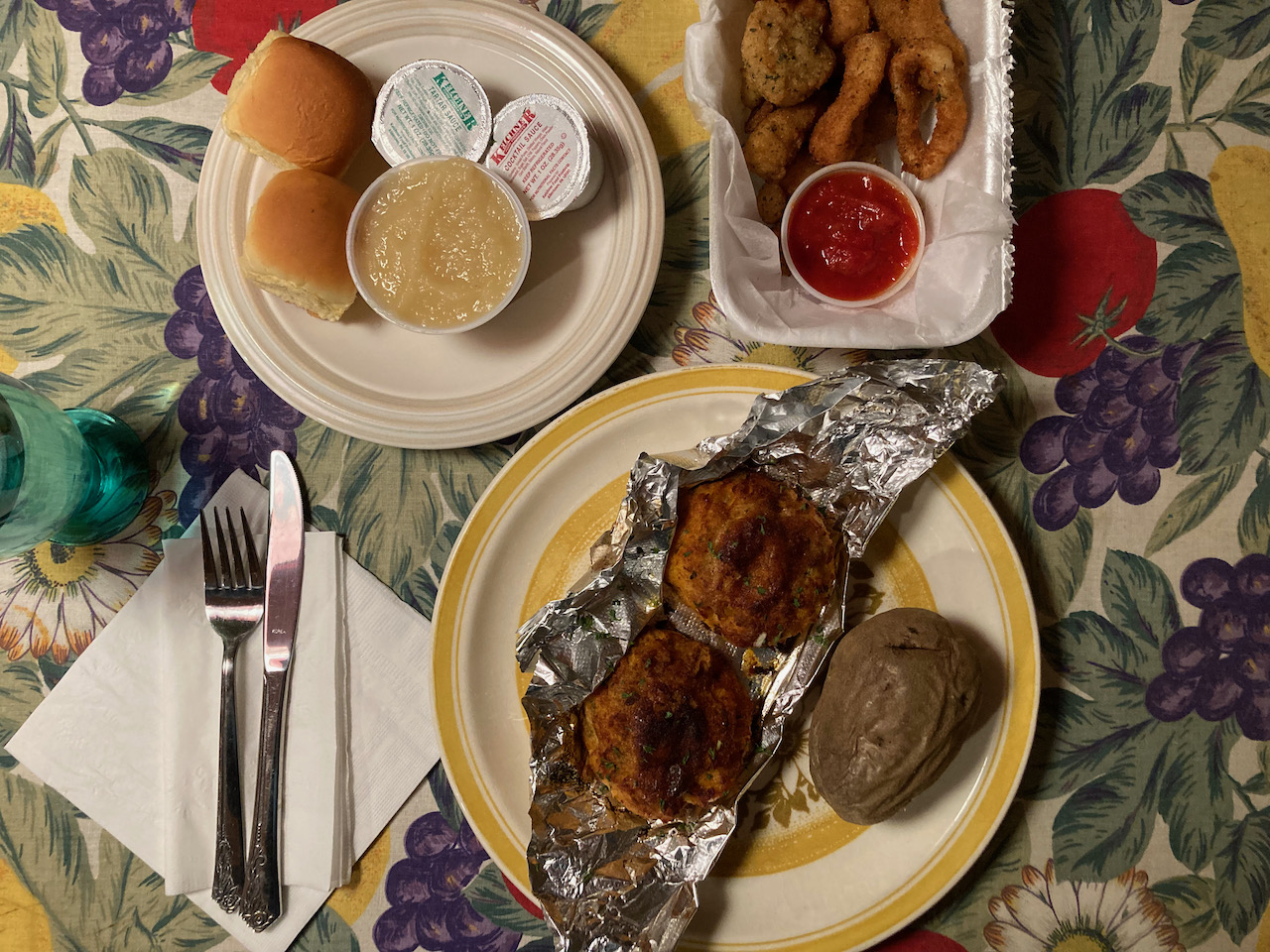 Two crab cakes on plate with baked potato. On another plate is apple sauce and rolls. In a styrofoam container is an order of calamari.