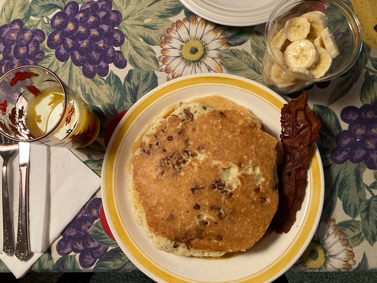 Pancakes on plate with an order of bacon. A glass of orange juice and a container of banana slices are also on the table.
