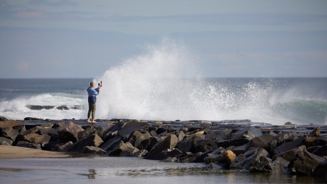 Man taking selfie on jetty, with rocks crashing in front of him.
