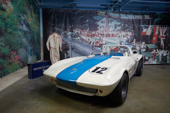 1963 Chevrolet Corvette Grand Sport (in blue and white) in front of photographic backdrop of Watkins Glen race track.