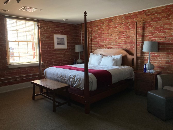 Hotel room with exposed brick walls. A four-poster king size bed is in the middle of the photo against the far wall.