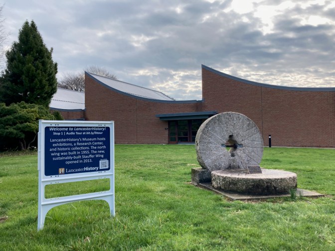 Lancaster history museum in background. In foreground, a sign says WELCOME TO LANCASTERHISTORY STOP 1 AUDIO TOUR.