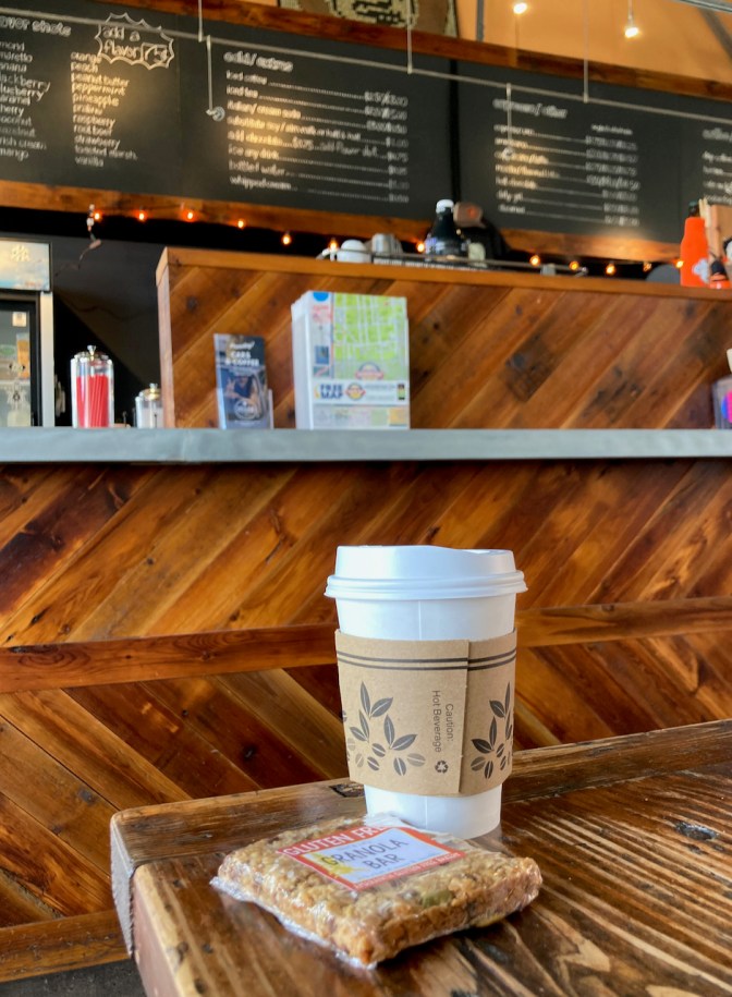 Cup of coffee and granola bar on table, with menu on wall behind counter in background.