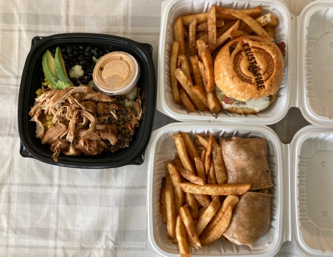 Three to-go containers. One holds a chicken and avocado rice bowl. Another holds a chicken wrap with french fries, and a third holds a burger, with the words CRUST & FIRE branded on the top of the bun.