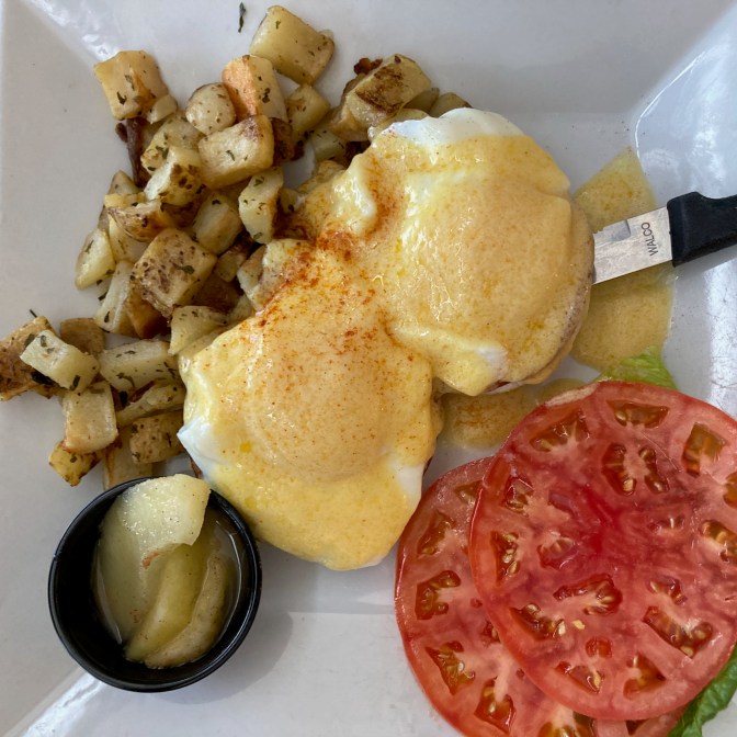 Eggs benedict on plate, with potatoes, tomato, and apple slices.