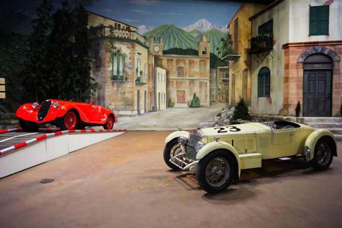 Two Alfa Romeos on display in front of diorama of Brescia for Mille Miglia race.