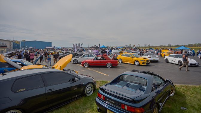 Wide-angle view of car show, with numerous classic and modified cars in parking lot.