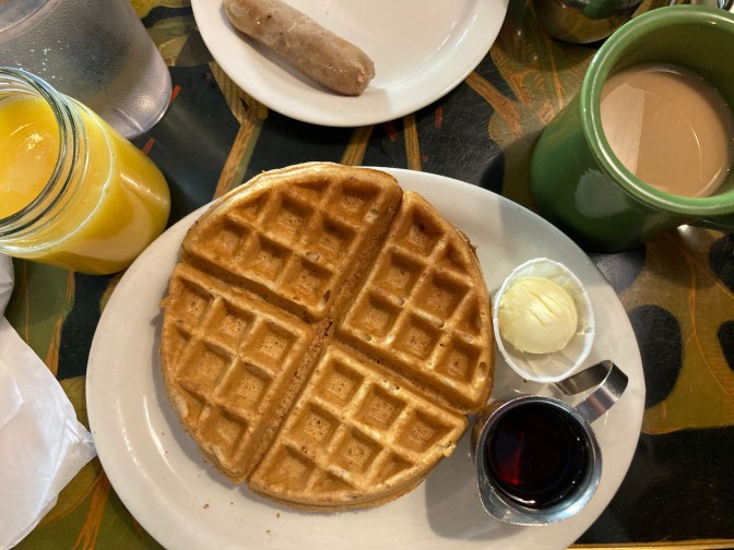 Waffle on plate, with sides of maple syrup and butter, a glass of orange juice, and a cup of coffee.