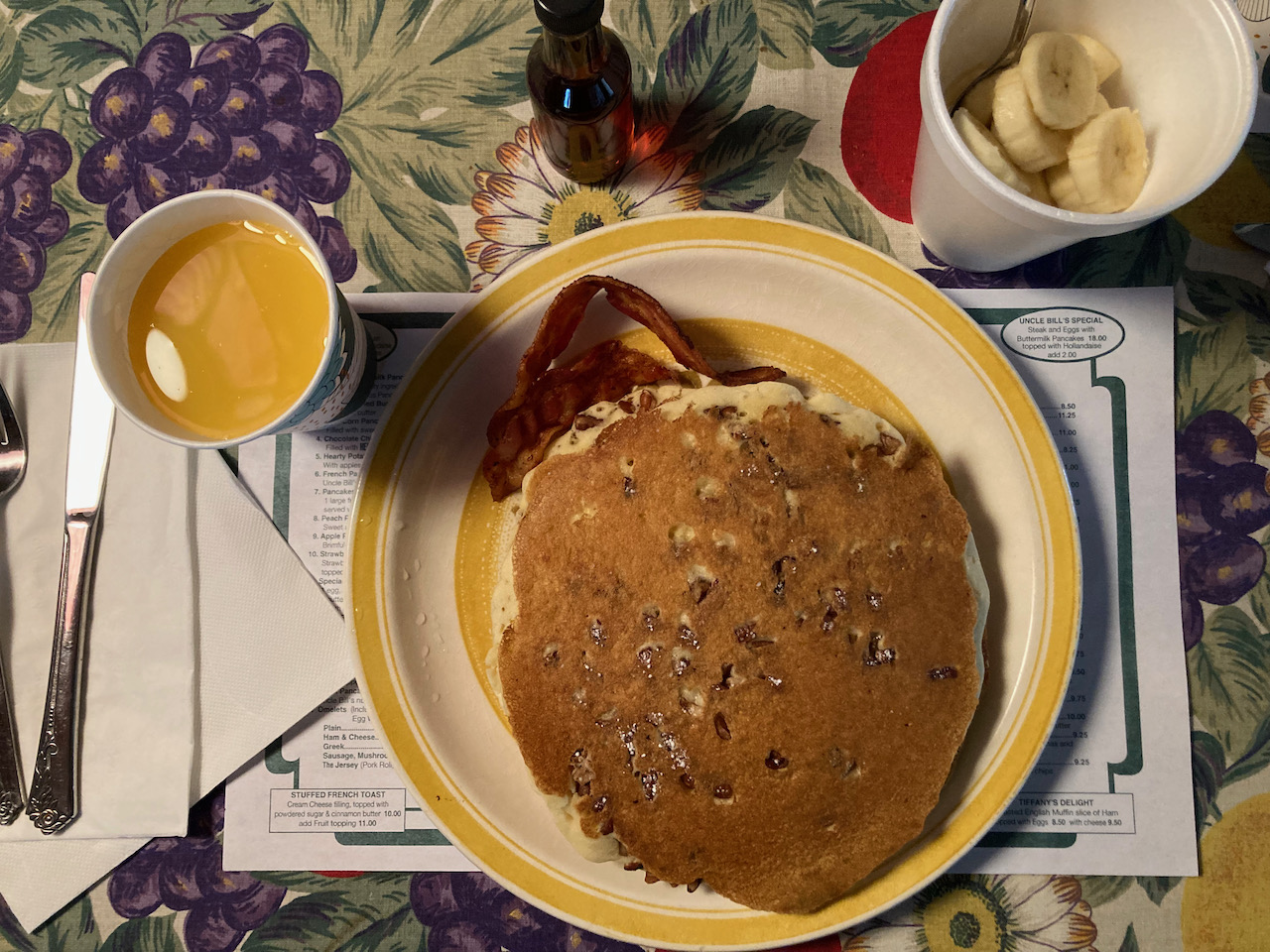 Pancakes and bacon on plate, with paper cup of orange juice and styrofoam cup of banana slices on table.