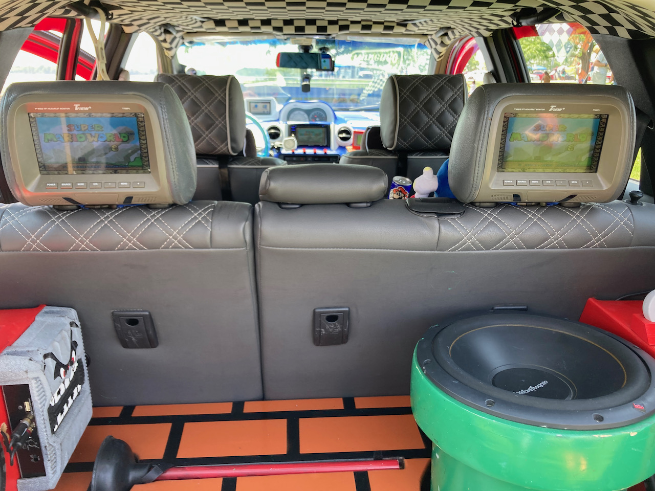 Trunk of Scion xA, with green pipe holding bass subwoofer, brick tiling, and seat rests with miniature TVs playing Super Mario World.
